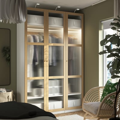 Licht eikenhouten PAX kledingkast met matglazen deuren, hoog tegen groene en beige wanden. Links: hanglamp, rechts: groot raam met groene stoel.