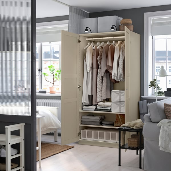 Open beige PAX kledingkast met kleding; Scharnierende deuren sluiten. Klein wit bed, doorschijnende gordijnen in de buurt.