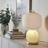 Een moderne PILBLIXT lamp met een matglazen kap op een metalen voetstuk. Hij staat naast boeken en een plant op een kast, wat het compacte formaat benadrukt.
