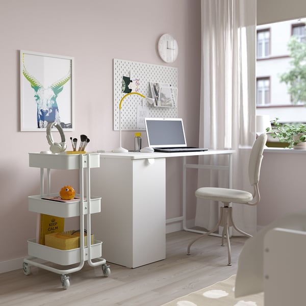 Compacte werkruimte met roze wanden met RÅSKOG trolley, laptop, koptelefoon, mok, containers en ophangbord. Opgefleurd door groot raam en witte stoel.
