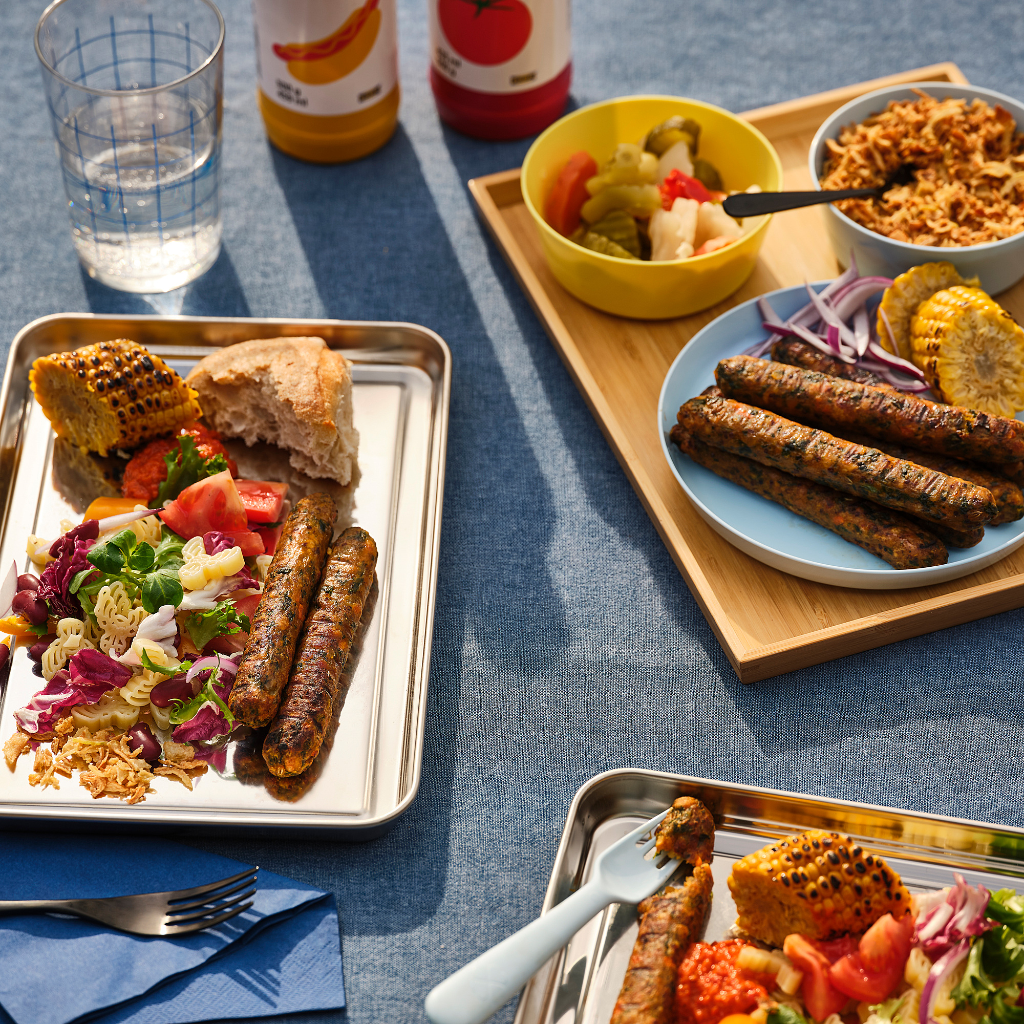 Vier borden met gegrilde worstjes, één bord met salade, brood en tomaten. Twee kommen met gemengde groenten en zuurkool, allemaal op een blauw tafelkleed.