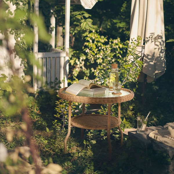 Ronde, rieten RISHOLMEN salontafel met glazen blad, met boek en drankje, gelegen in een groene tuin.