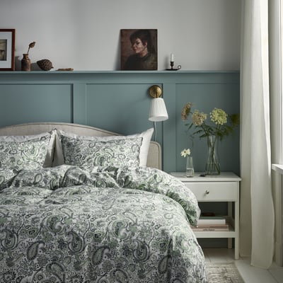 Slaapkamer met blauwgroene muur, paisley bed, nachtkastje met lamp en bloemen, portret op plank.