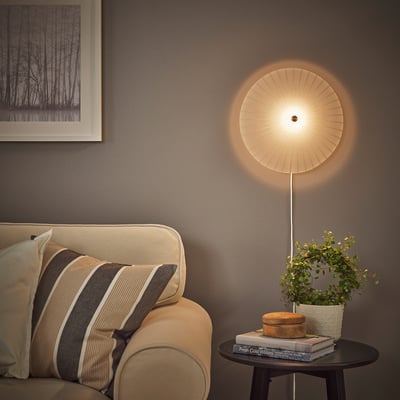 Ronde wandlamp op grijze muur naast foto, boven een bank met gestreept kussen en ronde tafel met boeken, doos en plant.