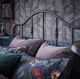 Slaapkamer met SAGSTUA zwart metalen bed, messing details, kleurrijke bloemenkussens, behang met donkere patronen.