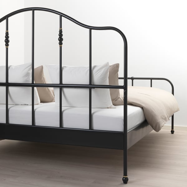 Zwart metalen bedframe met gebogen hoofdeinde en messingkleurige details, gekleed in wit linnen en kussens, geplaatst op een houten vloer.