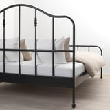 Zwart metalen bedframe met gebogen hoofdeinde en messingkleurige details, gekleed in wit linnen en kussens, geplaatst op een houten vloer.