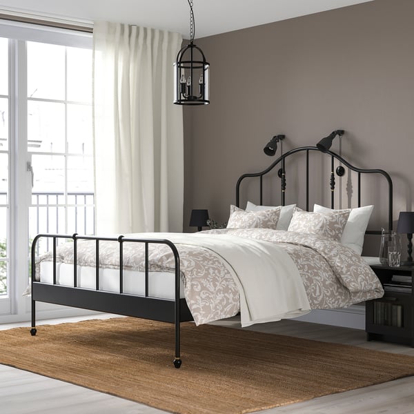 Gezellige slaapkamer met donker metalen SAGSTUA bed, messing details, keurig beddengoed, hanglamp, groot raam, nachtkastje, vloerkleed.