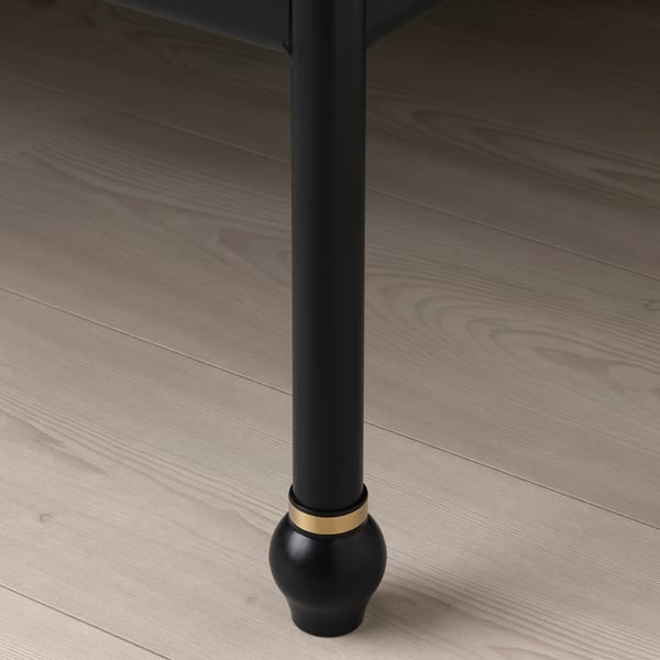 Close-up van SAGSTUA bed been, zwart met gouden accent, op houten vloer.
