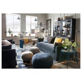 Moderne woonkamer met grijze bank, gele kussens, blauw vloerkleed, planten en grote ramen.