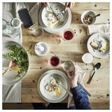 Een houten tafel met rustieke, aardse grijze steengoed borden, schalen en glazen. Mensen genieten van eten en drinken in een gezellige, informele eetomgeving.