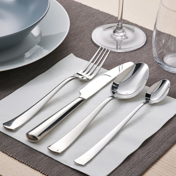 Gepolijst roestvrijstalen bestek op een servet, inclusief een mes, vork en lepel, gerangschikt op een tafel met borden en glazen.