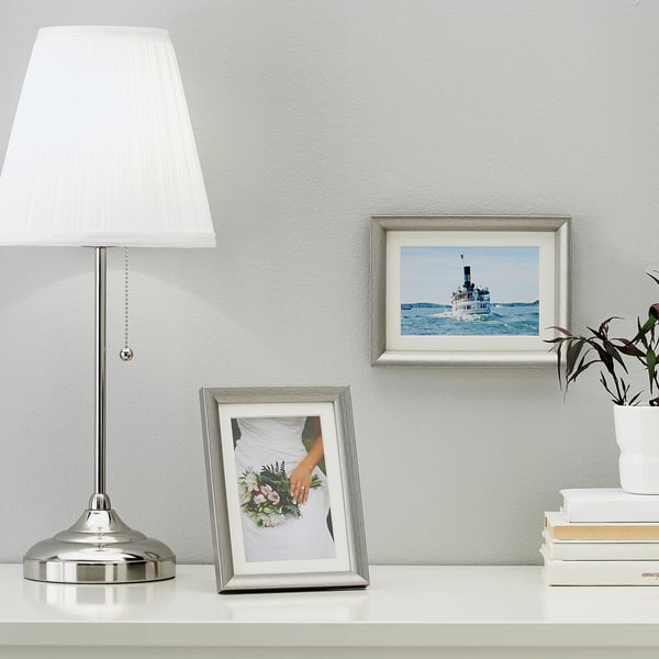 Een zilveren fotolijst op een tafel met een boeket, in de buurt van een lamp. Het frame kan staan of hangen.