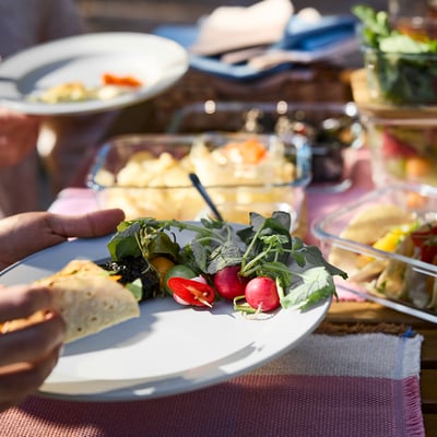 Hand houdt groen-grijzeSIMKRABBAplaat, gevuld met salade, radijs, en flatbread. Andere borden en eten in de buurt.