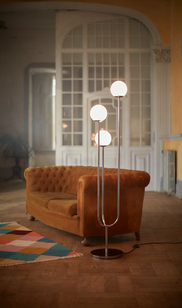 Moderne woonkamer met groot boograam, oranje gebogen bank, kleurrijk vloerkleed en SIMRISHAMN chromen vloerlamp met glazen bollen.