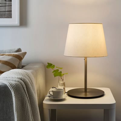 Een SKOTTORP lamp met beige kap op bijzettafel, omgeven door kopje, wijnbladeren en gezellige bank met witte deken. Zacht gloeiende plastic kap.