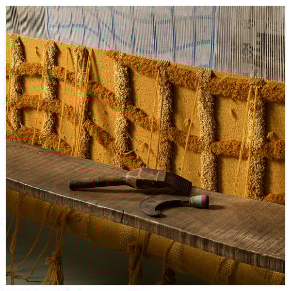 Handgeweven geelachtig bruin wollen tapijt met visnetpatroon op houten tafel. Bovenop rusten twee weefgereedschappen, die vakmanschap demonstreren.