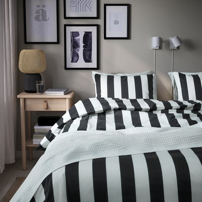 Een netjes opgemaakt bed met zwart-wit gestreept SLÅNHÖSTMAL beddengoed. De kamer heeft een moderne inrichting met ingelijste kunstwerken en een houten nachtkastje.