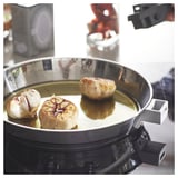 SLÄTROCKA pan met drie geroosterde knoflookbollen, zwart handvat, glanzend oppervlak op kookplaat.