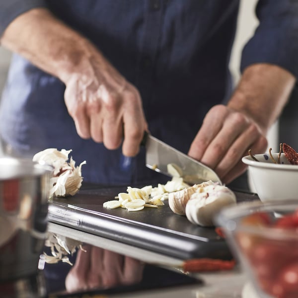Een persoon hakt knoflook op een snijplank met behulp van een mes. Het mes laat dunne plakjes achter, wat zorgt voor nauwkeurig snijden.