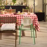 Zonnige keuken met roodgeruite tafelsetting, witte kinderstoel en potplanten.