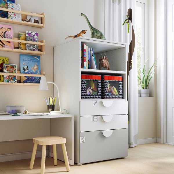 Kinderspeelkamer met witte SMÅSTAD boekenkast en opbergruimte, houten kruk, bureau, lamp, planken met boeken, speelgoed en kunstbenodigdheden.