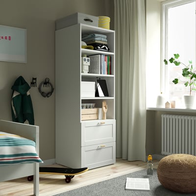 Grijs-witte SMÅSTAD boekenkast met boeken, kleding, decor, een klein bed en ingelijste kunst.
