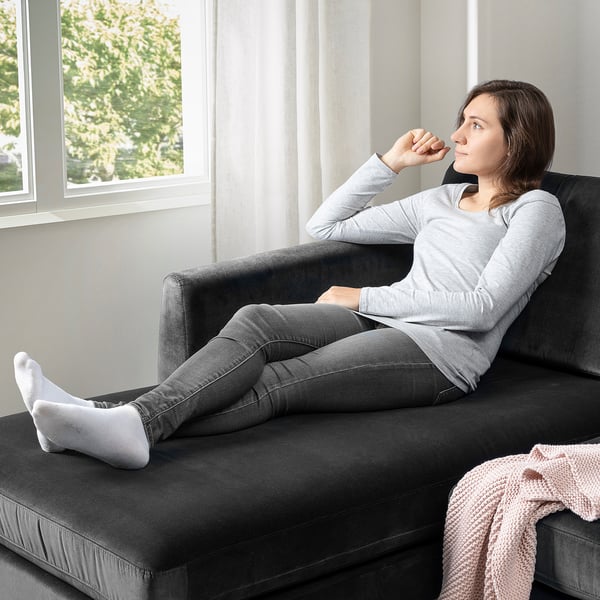 Een vrouw ontspant zich op een SMEDSTORP-bank bij een raam en geniet van de zachte, ondersteunende kussens die zijn ontworpen voor comfort en eenvoudige vloerreiniging.