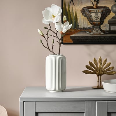 Kunstmatige witte magnolia in geribbelde beige vaas op dressoir, met decoratieve gouden pauw; Moderne home decor display.