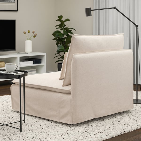 Een moderne, lichtgekleurde fauteuil met vierkante kussens op een wit vloerkleed naast een staande lamp en salontafel in een minimalistische woonkamer.