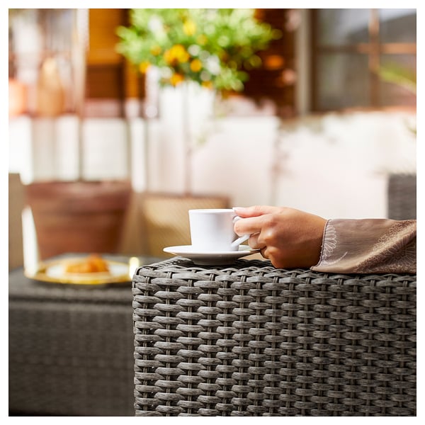 Een persoon die een witte koffiekop houdt aan een buitentafel met een gele plaat, bruine rieten meubels.