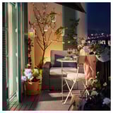 Gezellig balkon met beige SOLLERÖN tuinmeubilair, bloemen en wijn opstelling 's nachts.