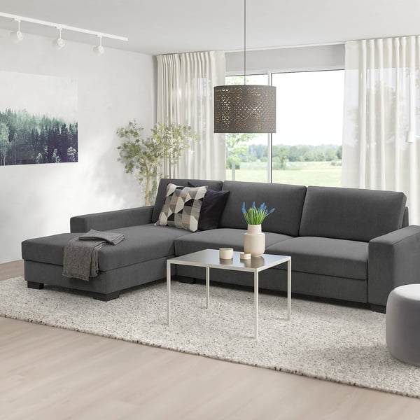 Moderne grijze L-vormige SÖRVALLEN bank, minimalistisch design. Gecentreerd onder een hanglamp, groter dan bijzettafel & plant, gezellig & ruim.