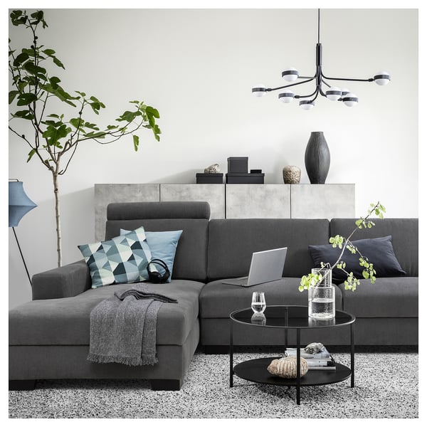 Moderne grijze sectionele bank met kussens met patronen, strakke salontafel, hanglampen en een grote plant in een minimalistische leefruimte.