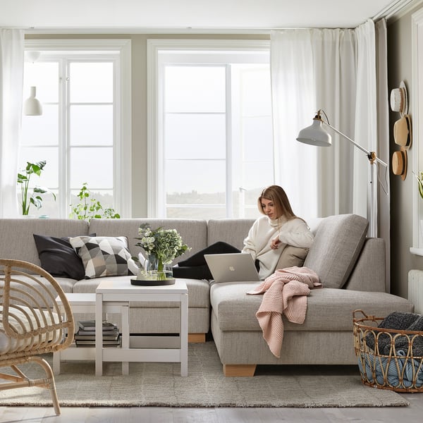 Moderne woonkamer SÖRVALLEN grijze bank met een vrouw die een laptop gebruikt, omgeven door natuurlijk licht, planten en een minimalistische inrichting.