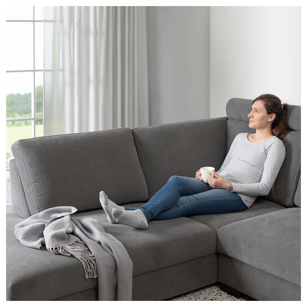 Vrouw ontspant zich op moderne grijze SÖRVALLEN-bank, met een mok, door een groot raam met doorschijnende gordijnen.