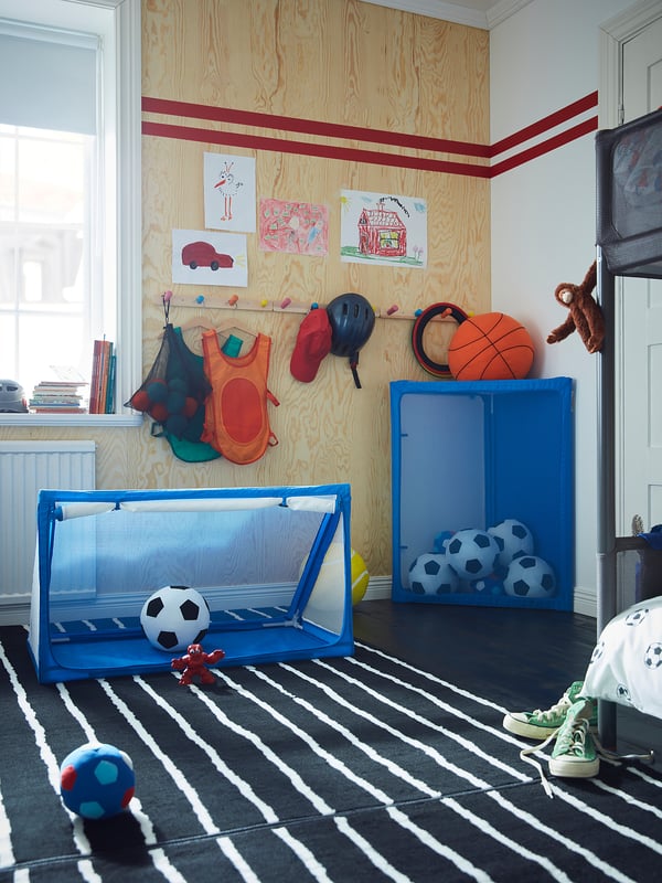 Speelkamer met blauwe sportuitrusting, gestreept tapijt, houten muur, kindertekeningen, speelgoed.