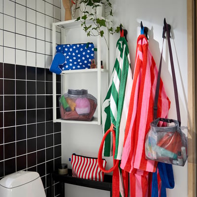 Badkamerscène met kleurrijke handdoeken en mesh tassen; zichtbaar: blauwe stippen, rood gestreepte tassen, hangend aan de muur; SPJUTSÖ tas formaat kleiner dan handdoeken.