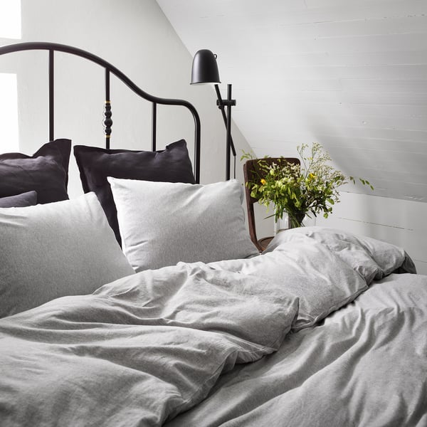 Onopgemaakt bed met grijs SPJUTVIAL dekbedovertrek, donkere kussens, wandlamp, bloemen.