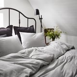 Onopgemaakt bed met grijs SPJUTVIAL dekbedovertrek, donkere kussens, wandlamp, bloemen.