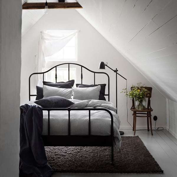 Gezellige slaapkamer met zwart metalen bed, witte muren, grijs beddengoed en een vaas met gele bloemen.