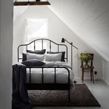 Gezellige slaapkamer met zwart metalen bed, witte muren, grijs beddengoed en een vaas met gele bloemen.