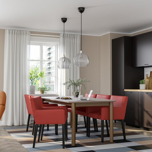 Moderne eethoek met STRANDTORP tafel en rode stoelen, twee glazen hanglampen, planten en geometrisch tapijt.