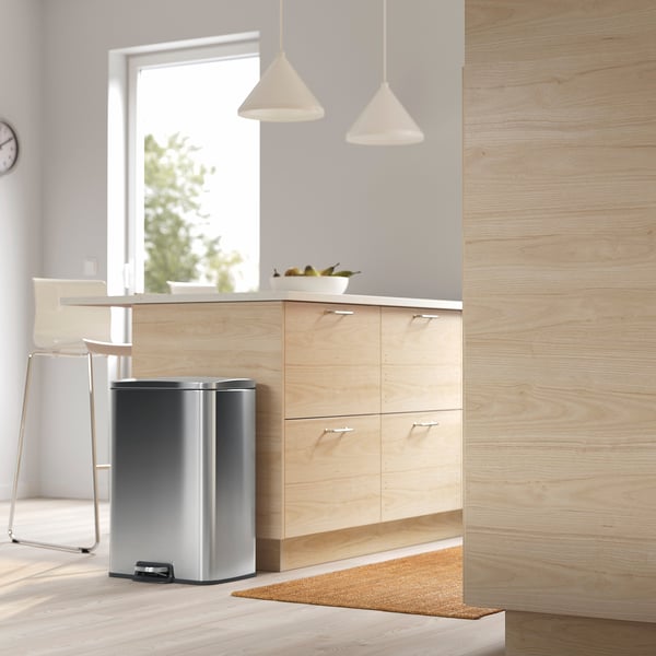 Moderne minimalistische keuken met een RVS pedaalemmer onder lichthouten kasten, hanglampen en een raam.
