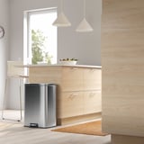 Moderne minimalistische keuken met een RVS pedaalemmer onder lichthouten kasten, hanglampen en een raam.
