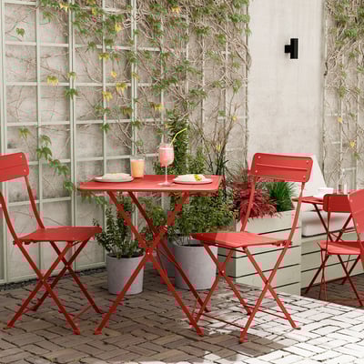 Felrode metalen patioset met klapstoelen en tafel. Perfect voor kleine ruimtes. Groene planten omringen.