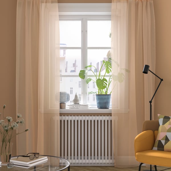 Lichte kamer met grote lichtoranje TERESIA gordijnen, een mosterdkleurige stoel en planten op een witte radiator onder een raam.