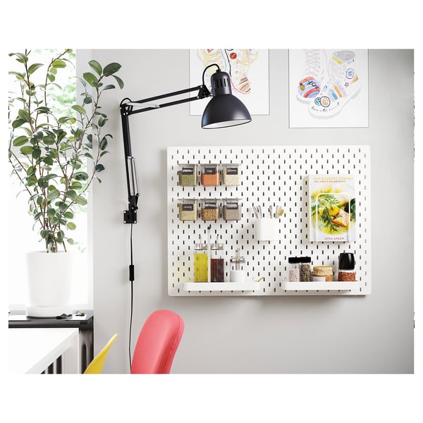 Een witte pegboard wandopstelling met een zwarte verstelbare bureaulamp, plant en kleurrijke stoel.