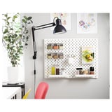 Een witte pegboard wandopstelling met een zwarte verstelbare bureaulamp, plant en kleurrijke stoel.