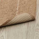 Hoek van beige jute tapijt op houten vloer, met getextureerde antislip achterkant.